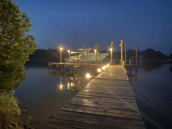 Long wooden dock at dusk, low dock lights and piling lights leading out to a boat on its lift